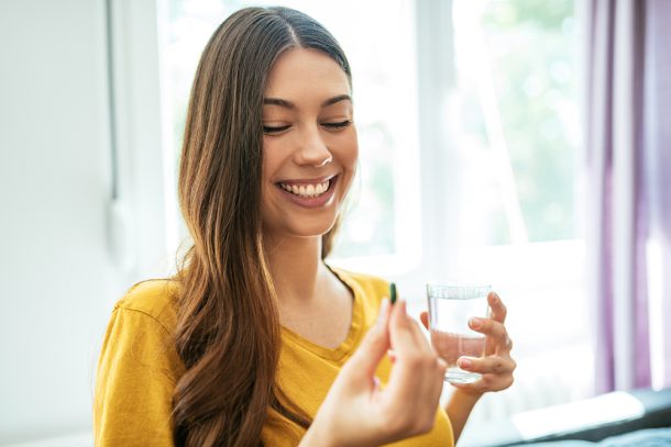 donna-assumendo-penicillina-a-casa Donna sorridente che assume una compressa di penicillina, insieme a un bicchiere d’acqua, nel salotto di casa