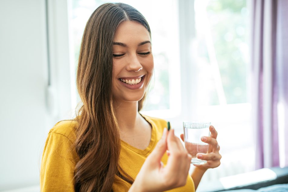 donna-assumendo-penicillina-a-casa Donna sorridente che assume una compressa di penicillina, insieme a un bicchiere d’acqua, nel salotto di casa