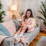 Giovane donna che idrata la pelle sul divano di casa per prevenire il prurito alle gambe con puntini rossi