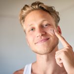 Giovane uomo che osserva davanti allo specchio l’infiammazione di un neo sul suo viso