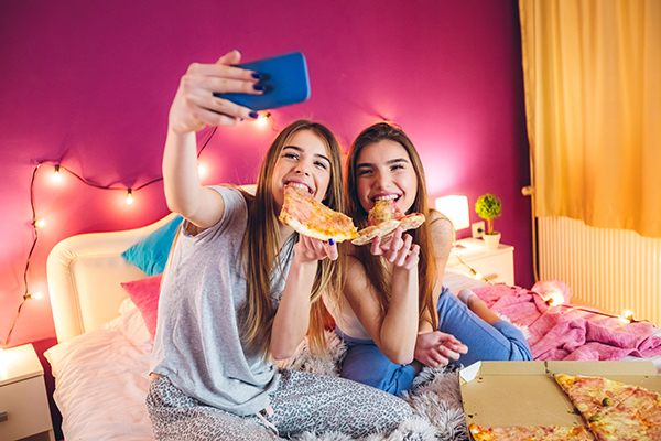 Amiche che condividono una pizza a letto durante il loro pigiama party, un’abitudine alimentare con possibile conseguenza di sviluppare l’obesità in giovane età
