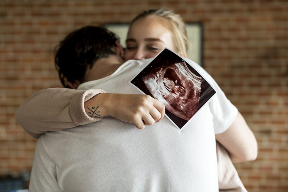 Coppia che si abbraccia emozionata, in cui la donna tiene in mano un’ecografia effettuata appena dopo i primi giorni dai sintomi della gravidanza