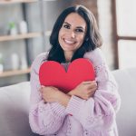 Donna sorridente seduta sul divano di casa che abbraccia una sagoma a forma di cuore, dopo avere approfondito cosa sono le coronarie