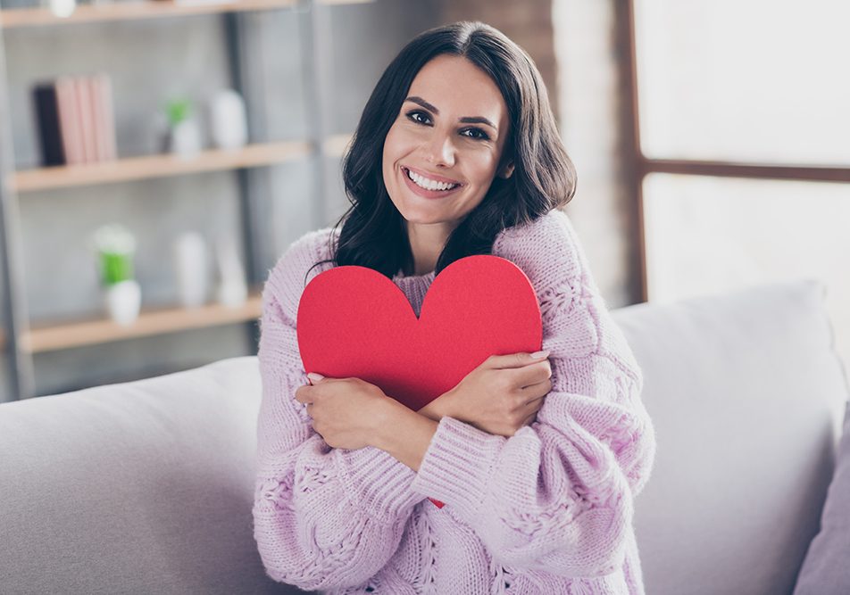 Donna sorridente seduta sul divano di casa che abbraccia una sagoma a forma di cuore, dopo avere approfondito cosa sono le coronarie