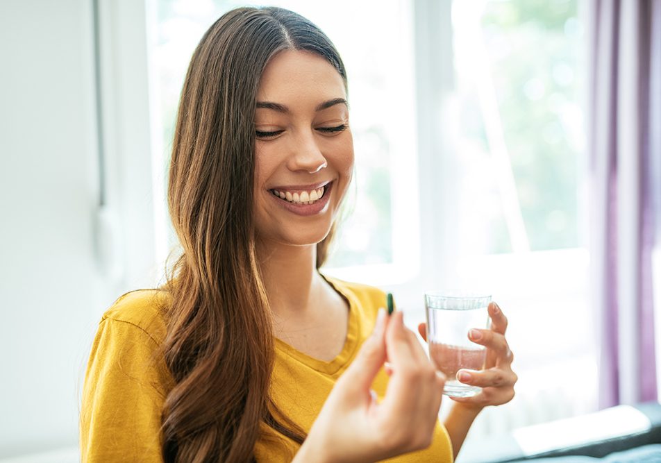 donna-assumendo-penicillina-a-casa Donna sorridente che assume una compressa di penicillina, insieme a un bicchiere d’acqua, nel salotto di casa