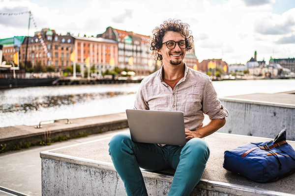 Giovane uomo che richiede l’esenzione ticket online attraverso il suo computer portatile mentre osserva il lungofiume della città