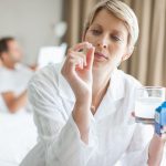 Donna seduta ai bordi del letto, su cui è presente anche il partner, che osserva una compressa di Oki, mentre tiene con l’altra mano un bicchiere d’acqua insieme alla scatola del farmaco
