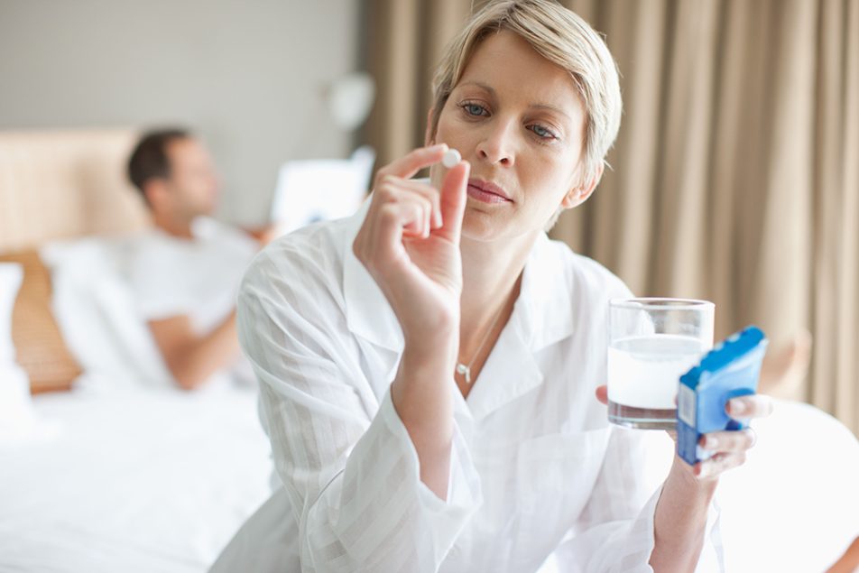 donna-a-letto-osservando-compressa-oki Donna seduta ai bordi del letto, su cui è presente anche il partner, che osserva una compressa di Oki, mentre tiene con l’altra mano un bicchiere d’acqua insieme alla scatola del farmaco