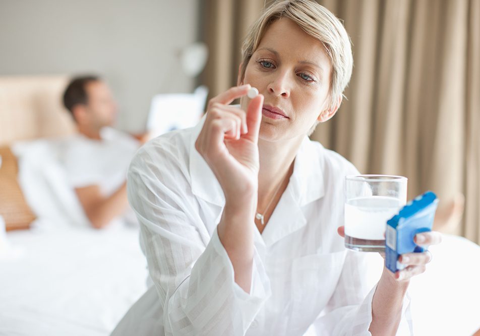 donna-a-letto-osservando-compressa-oki Donna seduta ai bordi del letto, su cui è presente anche il partner, che osserva una compressa di Oki, mentre tiene con l’altra mano un bicchiere d’acqua insieme alla scatola del farmaco