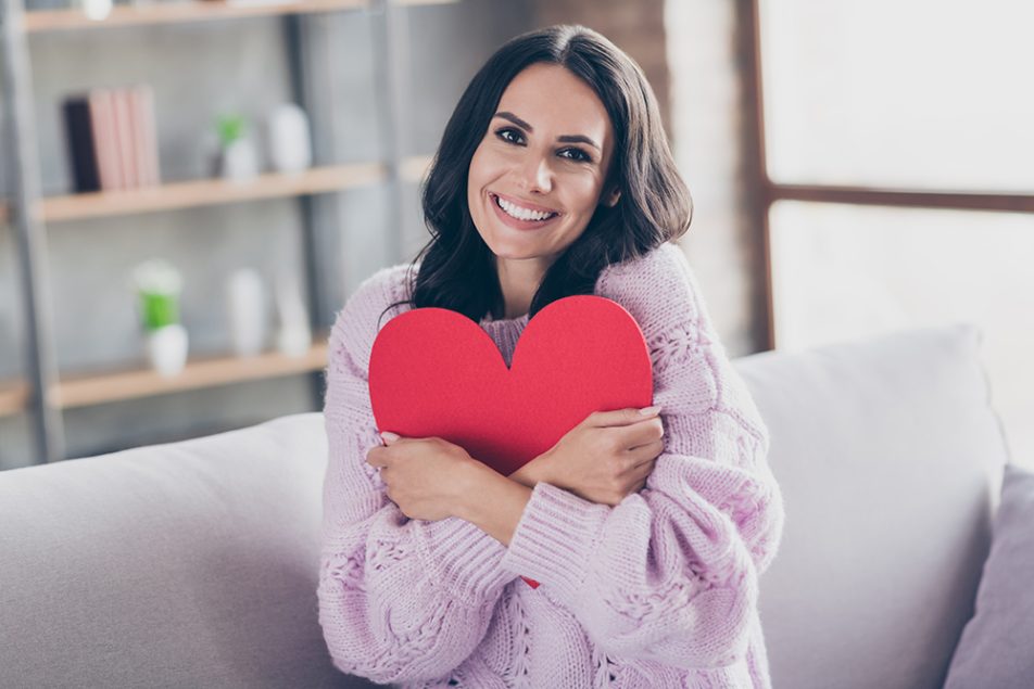 Donna sorridente seduta sul divano di casa che abbraccia una sagoma a forma di cuore, dopo avere approfondito cosa sono le coronarie