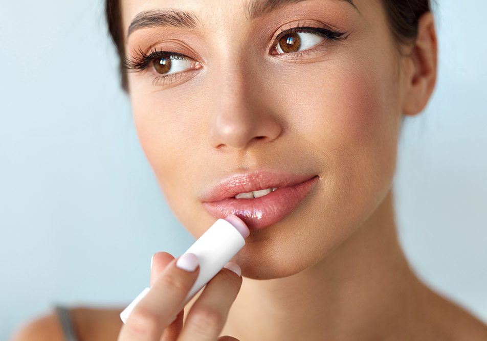 Donna mora che esamina il modo in cui ottenere delle belle labbra carnose applicando il balsamo labbra di fronte allo specchio