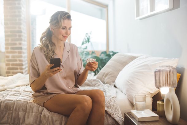 donna-in-pigiama-seduta-a-letto-assumendo-femara-e-consultando-internet-dallo-smartphone Donna in pigiama seduta a letto che assume una compressa di Femara e che consulta internet dal suo smartphone