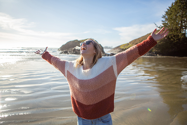 Giovane donna in spiaggia che alza le braccia con entusiasmo essendo grata della riduzione del tumore al pancreas di cui è affetta