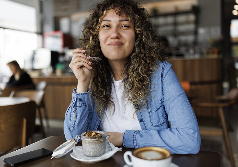 Giovane donna sorridente che mangia un budino con semi di chia per colazione seduta al tavolino di un bar