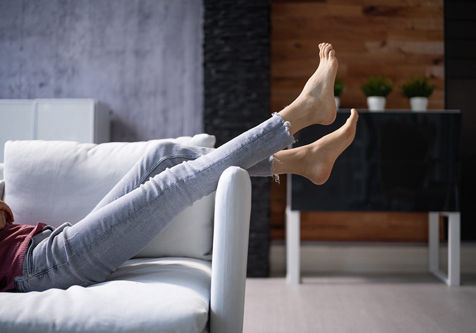 donna-sdraiandosi-sul-divano-sollevando-piedi-gonfi Donna sdraiata supina sul divano con le gambe alzate per fornire sollievo ai piedi gonfi