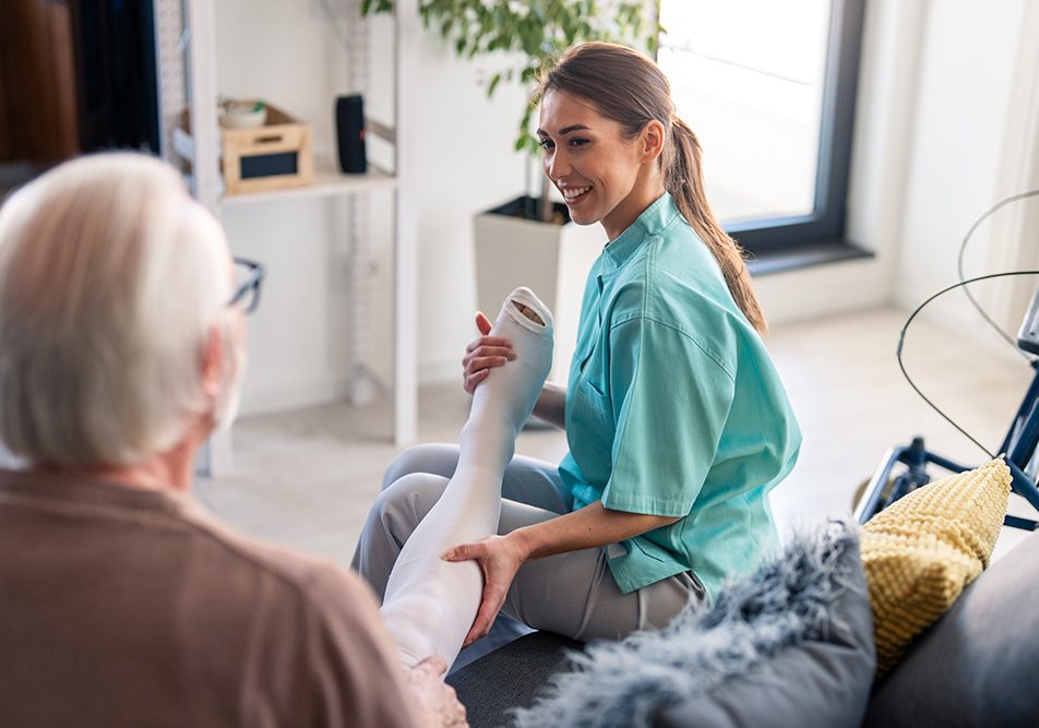 infermiera-assistendo-uomo-anziano-con-calze-compressive Infermiera sorridente che assiste un uomo anziano in salotto per indossare delle calze a compressione