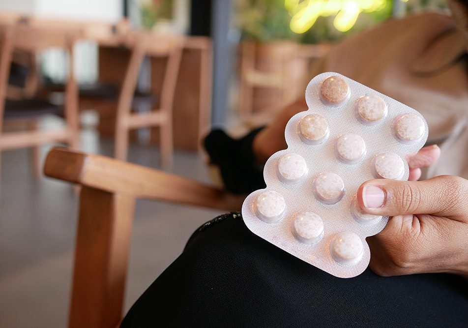 Mano che afferra un blister di compresse Oki all’interno di un bistrot accogliente con tavoli e sedie in legno