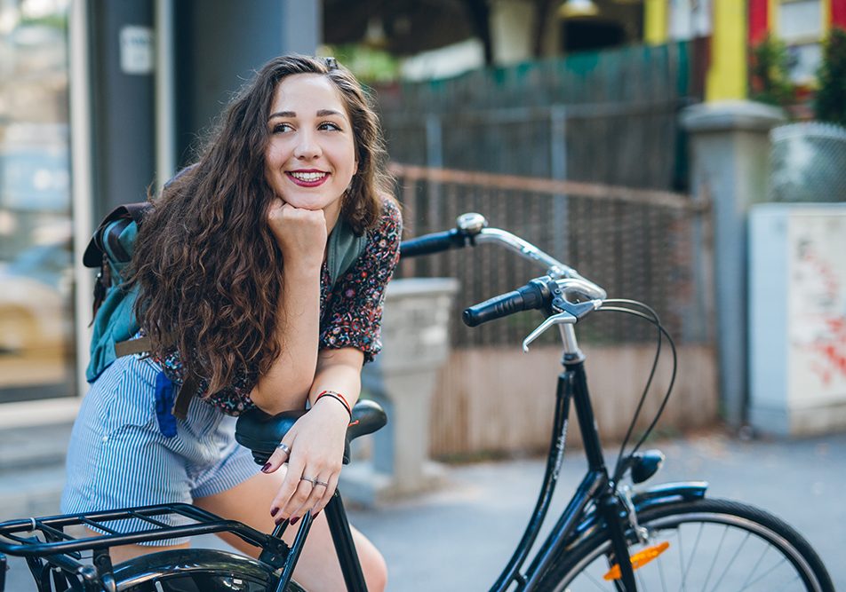 Ragazza in sosta lungo una via cittadina che si appoggia al sellino della sua bicicletta riflettendo se salire o meno per via dei dolori da ciclo senza ciclo