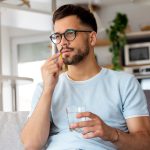 Giovane uomo che assume un farmaco antinfiammatorio mentre è seduto sul divano di casa, dopo essersi informato su quanto dura l’effetto antidolorifico dell’Oki