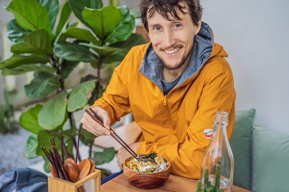 Uomo seduto ad un tavolo accanto ad una pianta, che mangia una bowl di riso e salmone, integrando l’Omega 3 e i benefici di tale nutriente attraverso un’alimentazione sana
