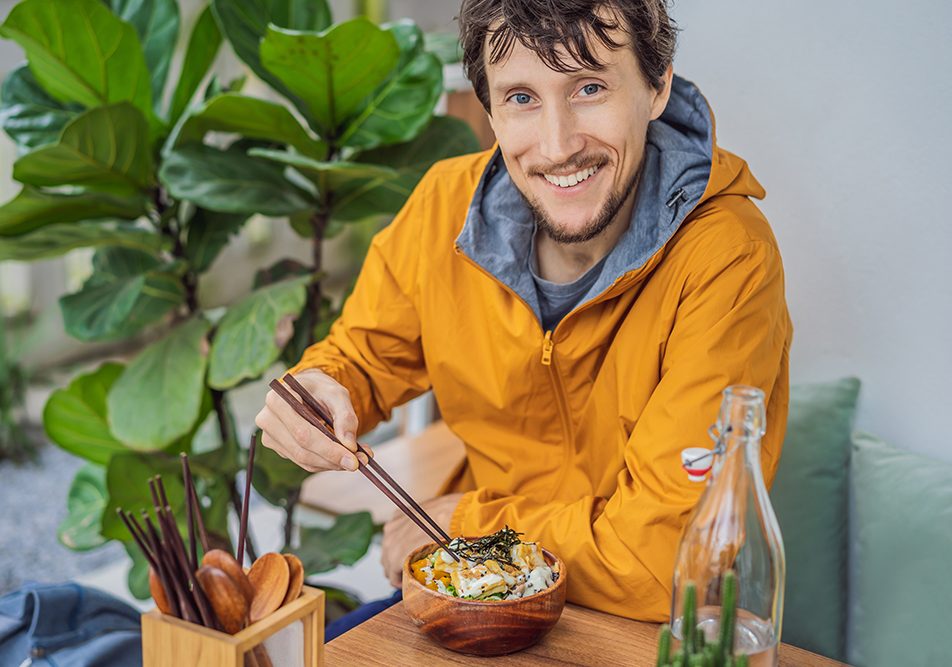 uomo-mangiando-bowl-con-salmone-omega-3-benefici Uomo seduto ad un tavolo accanto ad una pianta, che mangia una bowl di riso e salmone, integrando l’Omega 3 e i benefici di tale nutriente attraverso un’alimentazione sana