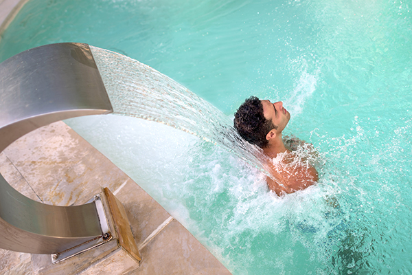 Uomo all’interno di una piscina di acqua sulfurea, al di sotto di una cascata con getto utile a combattere i dolori da cervicale