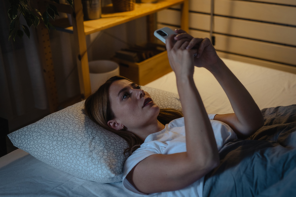 Donna a letto che consulta lo smartphone per lungo tempo prima di addormentarsi, senza considerare di quante ore abbia bisogno di dormire