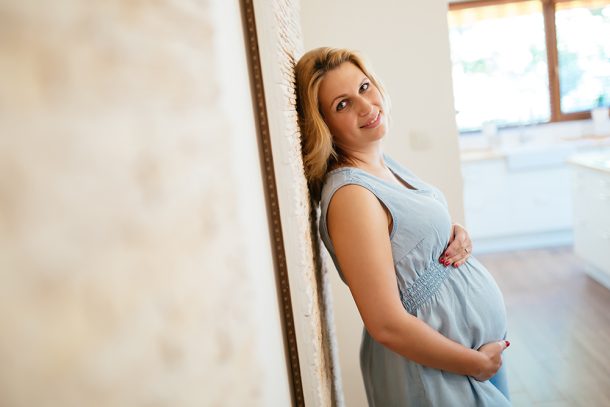 donna-affrontando-cisti-ovarica-in-gravidanza Donna incinta sorridente appoggiata a una parete mentre si tocca il pancione, che sta affrontando i disturbi della cisti ovarica in gravidanza
