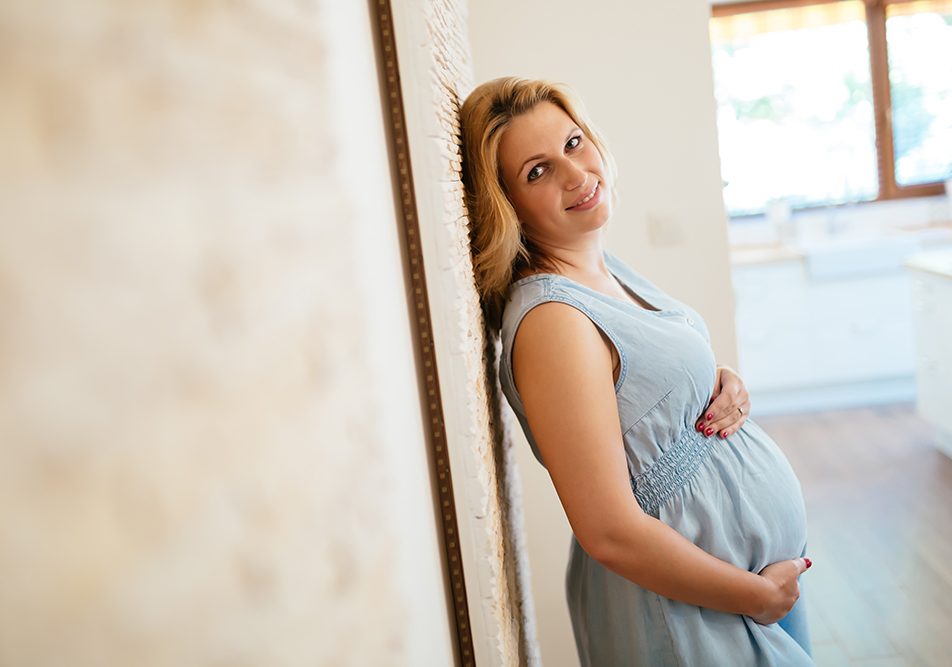donna-affrontando-cisti-ovarica-in-gravidanza Donna incinta sorridente appoggiata a una parete mentre si tocca il pancione, che sta affrontando i disturbi della cisti ovarica in gravidanza