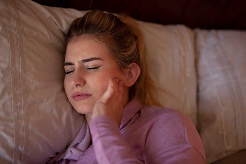 donna-con-bruxismo-sintomi-toccandosi-mandibola Donna a letto con sintomi del bruxismo mentre si tocca la mandibola dolorante