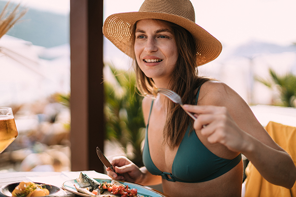 Donna che, durante le vacanze estive, mangia pesce alla griglia presso un bar sulla spiaggia, essendo a conoscenza dei cibi in cui trovare la vitamina D