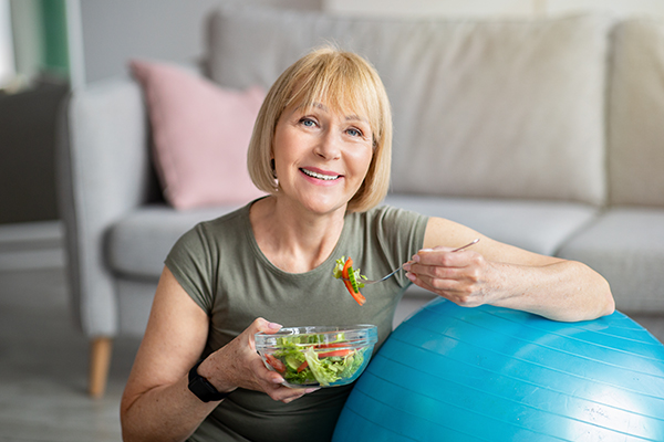 Donna senior che pratica esercizio fisico in casa con una fitball e mangia un’insalata sana e leggera. prevenendo l’insorgere dell’ictus