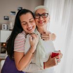 In un accogliente salotto, figlia adulta che abbraccia affettuosamente la madre affetta da tumore che indossa un paio di occhiali e un foulard sul capo