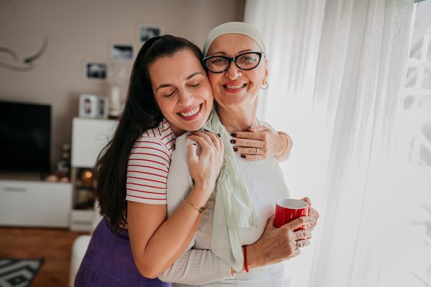 In un accogliente salotto, figlia adulta che abbraccia affettuosamente la madre affetta da tumore che indossa un paio di occhiali e un foulard sul capo