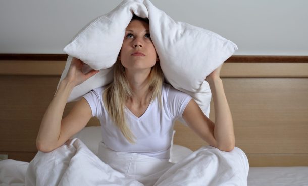 giovane-donna-che-soffre-di-disturbi-del-sonno Giovane donna seduta sul letto con cuscino sulla testa che soffre di disturbi del sonno
