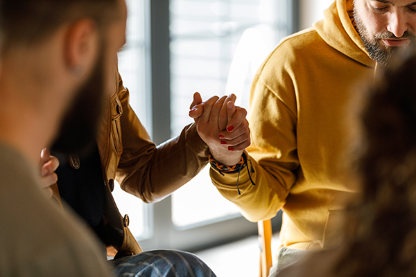 Gruppo di persone adulte sedute in cerchio durante una sessione di psicoterapia, tenendosi per mano per affrontare le sfide della convivenza con il tumore