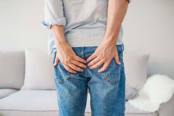 uomo-con-cisti-pilonidale-scoppiata-tenendo-mani-sulle-natiche Uomo in jeans con una cisti pilonidale scoppiata che tiene le mani sulle natiche per attenuare il dolore
