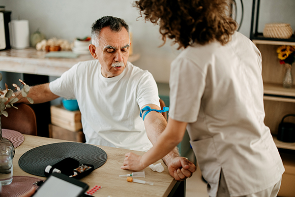 Uomo maturo in ambiente domestico insieme all’infermiera, che si sottopone agli esami del sangue per verificare l’andamento della neoplasia prostatica