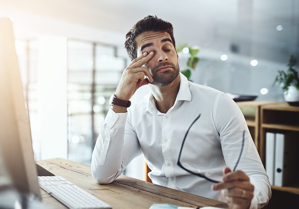 Uomo in ufficio seduto davanti al computer, che si sfrega gli occhi e si interroga sulle possibili cause della sua costante sonnolenza