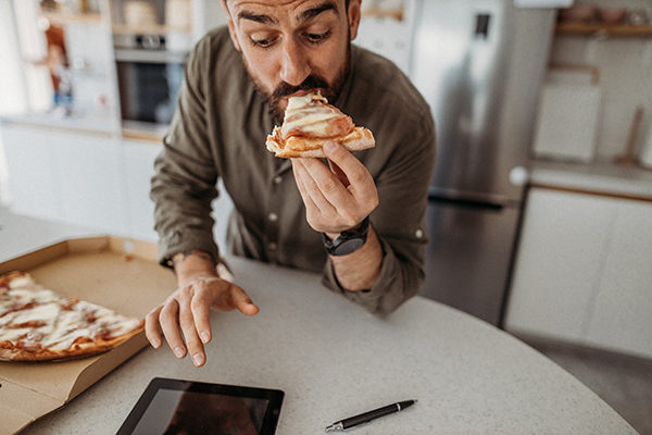 Uomo in cucina che, mentre consulta il tablet, mangia velocemente una pizza farcita ricca di calorie in eccesso, una delle cause della trigliceridemia alta