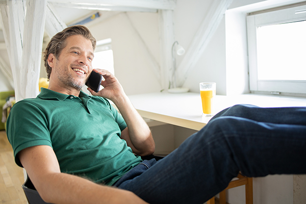 Uomo maturo a lavoro da casa che migliora la circolazione alle gambe sollevandole sulla scrivania mentre conversa al telefono