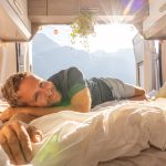 Uomo che si rilassa sul letto del camper con vista sulle montagne, esplorando i metodi per riuscire a dormire bene durante il suo viaggio