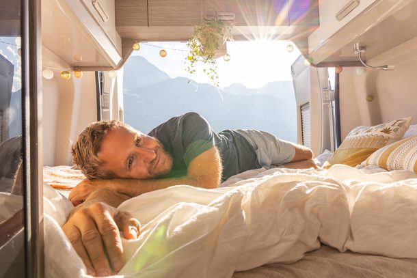 uomo-rilassandosi-in-camper-esplorando-metodi-per-dormire-bene Uomo che si rilassa sul letto del camper con vista sulle montagne, esplorando i metodi per riuscire a dormire bene durante il suo viaggio