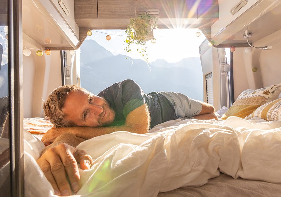 uomo-rilassandosi-in-camper-esplorando-metodi-per-dormire-bene Uomo che si rilassa sul letto del camper con vista sulle montagne, esplorando i metodi per riuscire a dormire bene durante il suo viaggio
