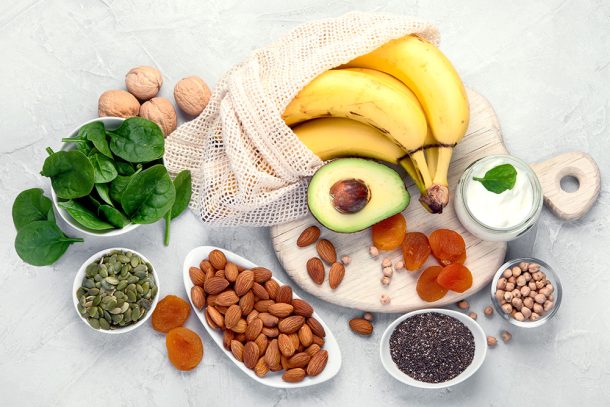 alimenti-ricchi-di-manesio-e-potassio-tra-cui-frutta-secca-cereali-e-banane Tavola con un tagliere e una serie di piattini contenenti alimenti ricchi di magnesio e potassio, tra cui frutta secca, cereali e banane
