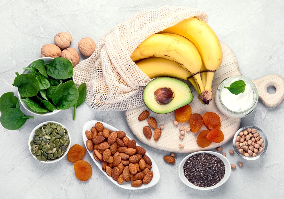 Tavola con un tagliere e una serie di piattini contenenti alimenti ricchi di magnesio e potassio, tra cui frutta secca, cereali e banane