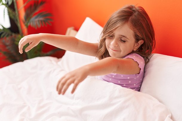 bambina-seduta-a-letto-con-disturbi-del-sonno-sonnambulismo-alzando-braccia Bambina sorridente seduta sul letto che soffre di sonnambulismo, un disturbo del sonno comune in età infantile, e tiene le braccia alzate
