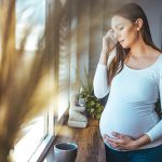 Giovane donna incinta con mal di testa in gravidanza che cerca sollievo guardando fuori dalla finestra del suo appartamento