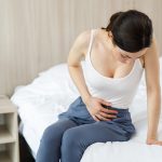 Giovane donna con sintomi del tumore alle ovaie seduta a letto che tocca il basso ventre dolorante con la mano
