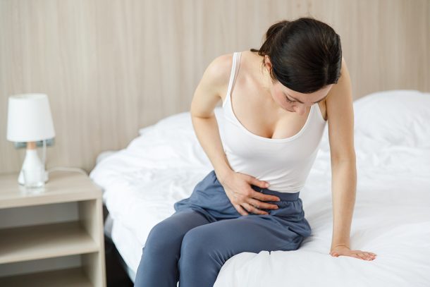 donna-con-tumore-ovaie-sintomi-toccando-basso-ventre-dolorante Giovane donna con sintomi del tumore alle ovaie seduta a letto che tocca il basso ventre dolorante con la mano